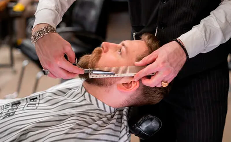 Homem de barba sentado inclinado e barbeiro com pente e tesoura fazendo barba do homem sentado