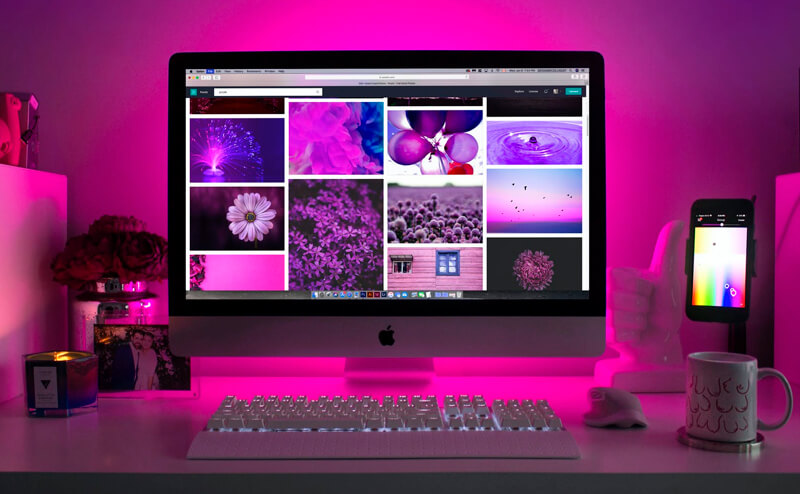 Escrivaninha com uma luz rosa, monitor da Apple e celular ao lado do iMAC. Na tela do monitor mostram fotos diversas.