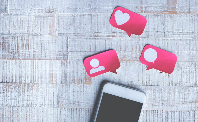 Ícones que representam as redes sociais. Cortado em cartolina na cor rosa, com ícone de usuário, coração e balão de mensagem. Smartphone abaixo dos ícones.